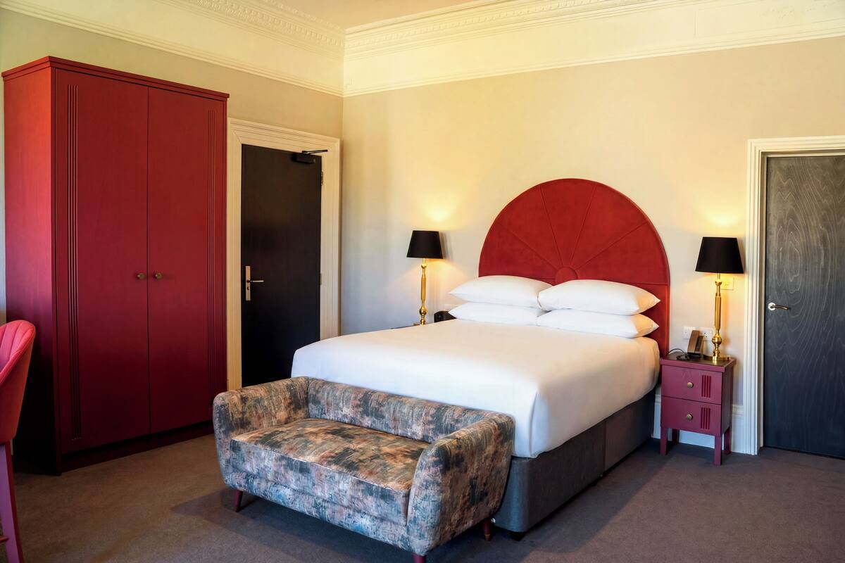 A bedroom with a bed, two bedside tables with lamps, a red wardrobe, and a patterned armchair.