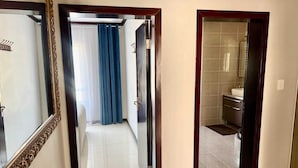 Un couloir mène à une salle de bain dont le sol est carrelé, qui est dotée d’un rideau de douche bleu et d’un miroir orné d’un cadre travaillé.