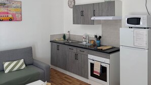 A modern kitchen with grey cabinets, a white refrigerator, and a microwave. There's a wooden floor, a grey sofa, and a white coffee table with a book and a tissue box.