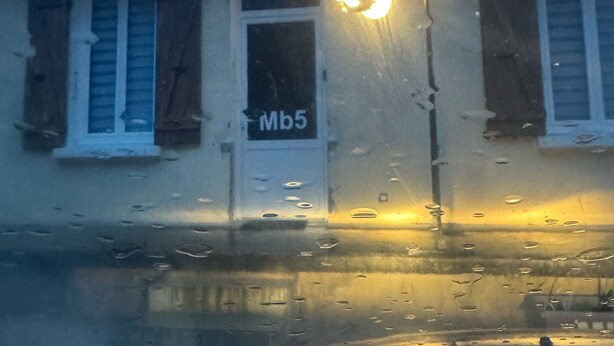 A building with a sign reading "Mb5" on the door, a chimney, and a window with shutters.