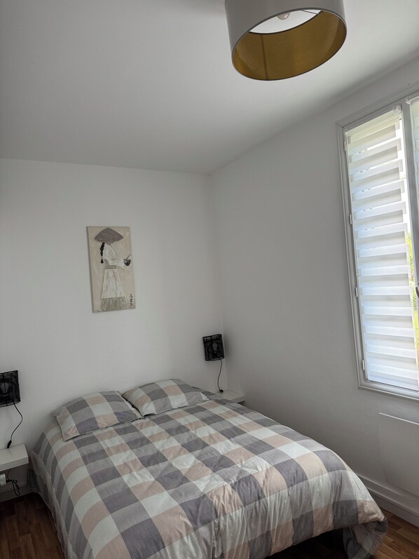 Une chambre à coucher avec un lit, un ventilateur fixé au mur, une fenêtre avec des stores et un tableau accroché au mur.