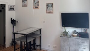 A modern living room with a wooden floor, a dining table with chairs, a TV mounted on the wall, and a grey cabinet with a plant.