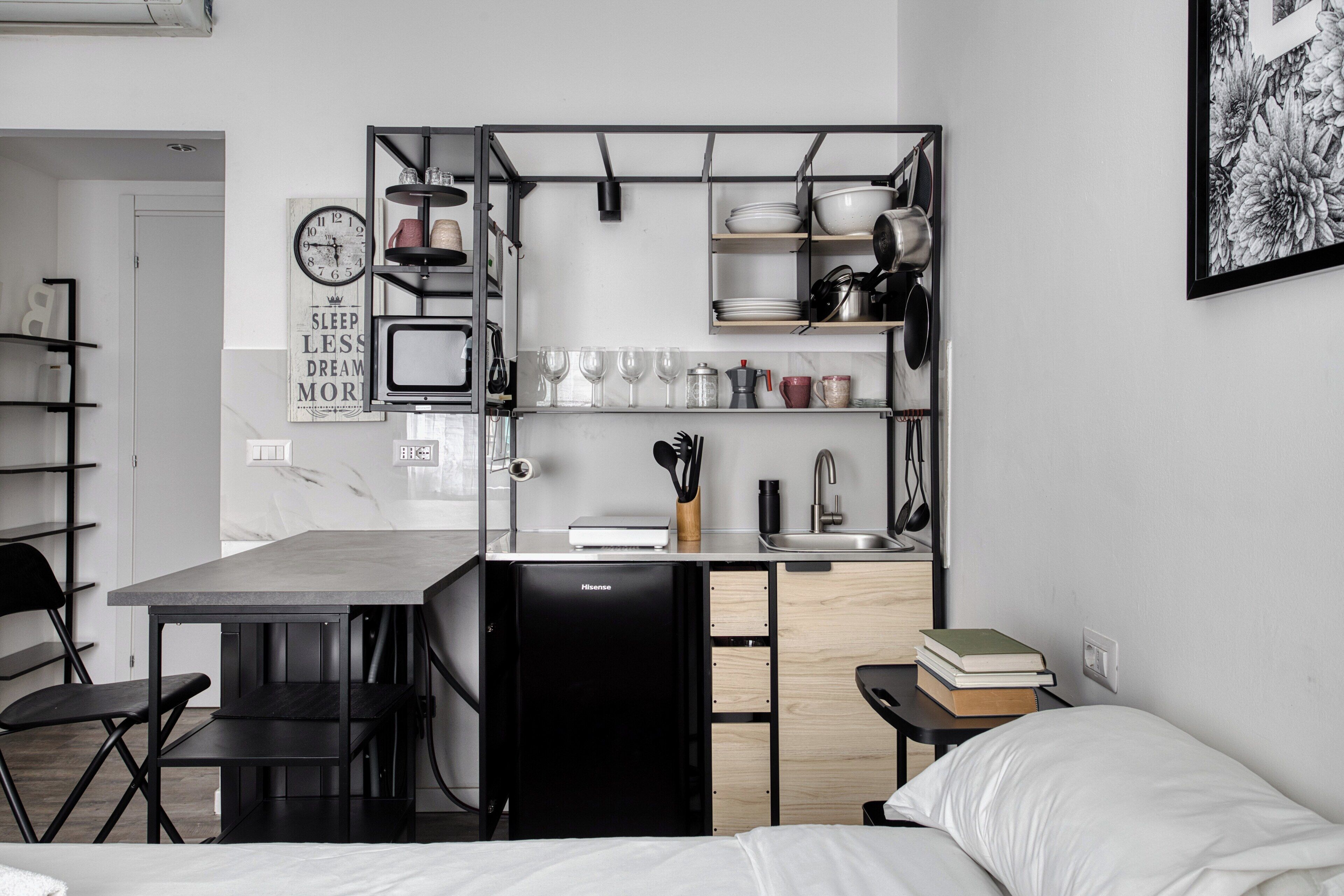 A compact kitchen with a dining table, a black refrigerator, and a sink. There are shelves above the counter holding dishes and glasses. A clock and a framed quote are on the wall.