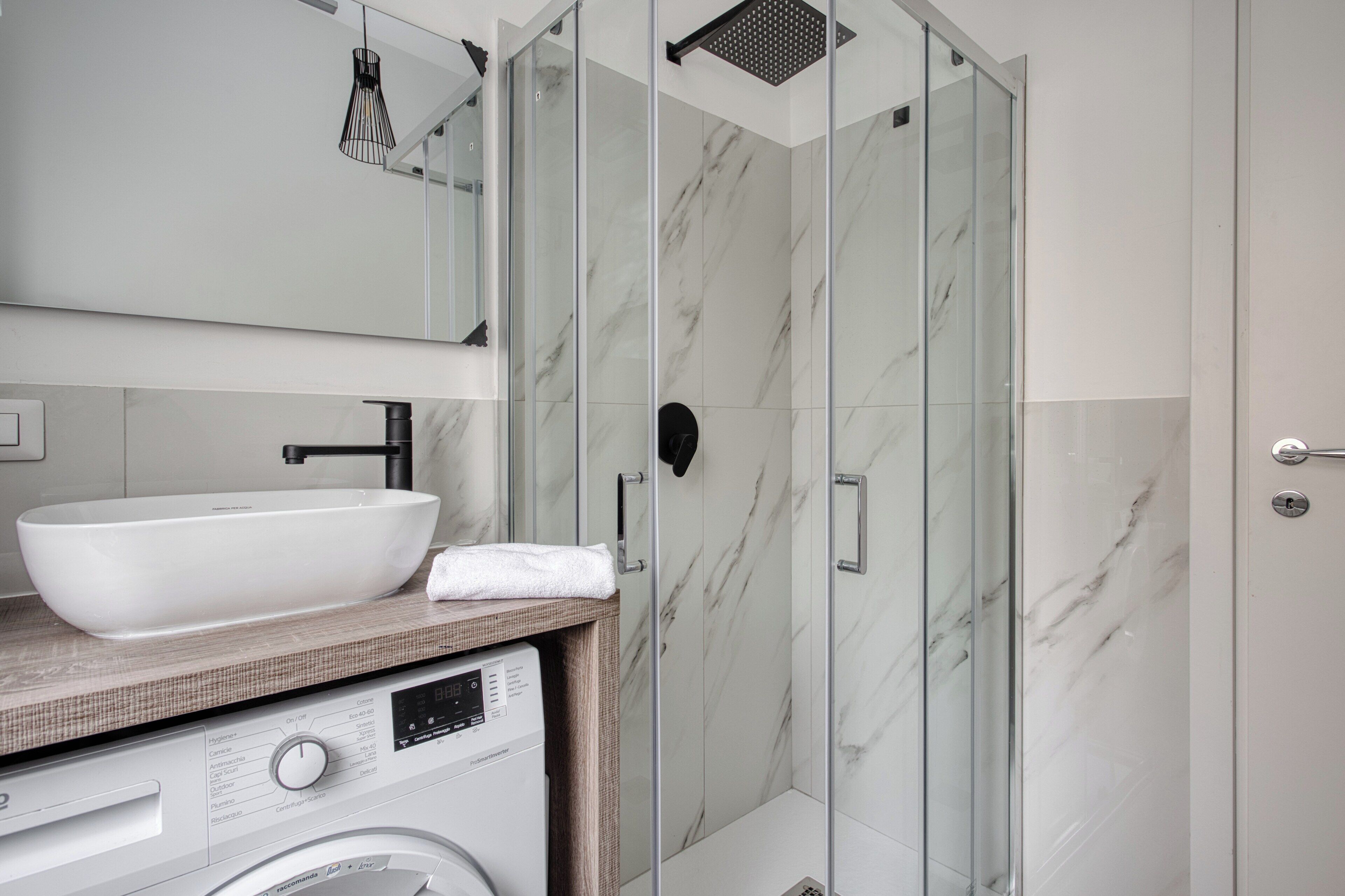 A modern bathroom with a large white sink, a glass shower enclosure, and a built-in washing machine.