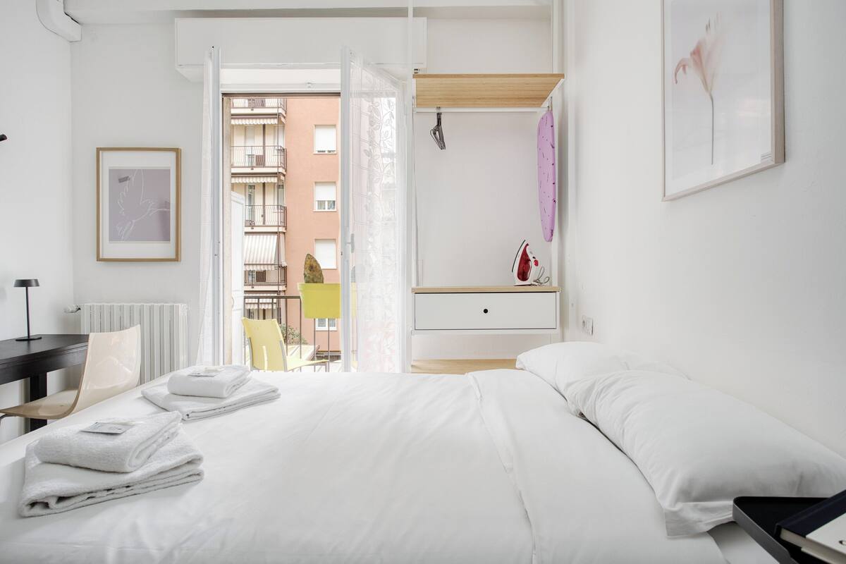 A hotel room with a bed, a bedside table, a chair, a window with a view of buildings, and a framed picture on the wall.