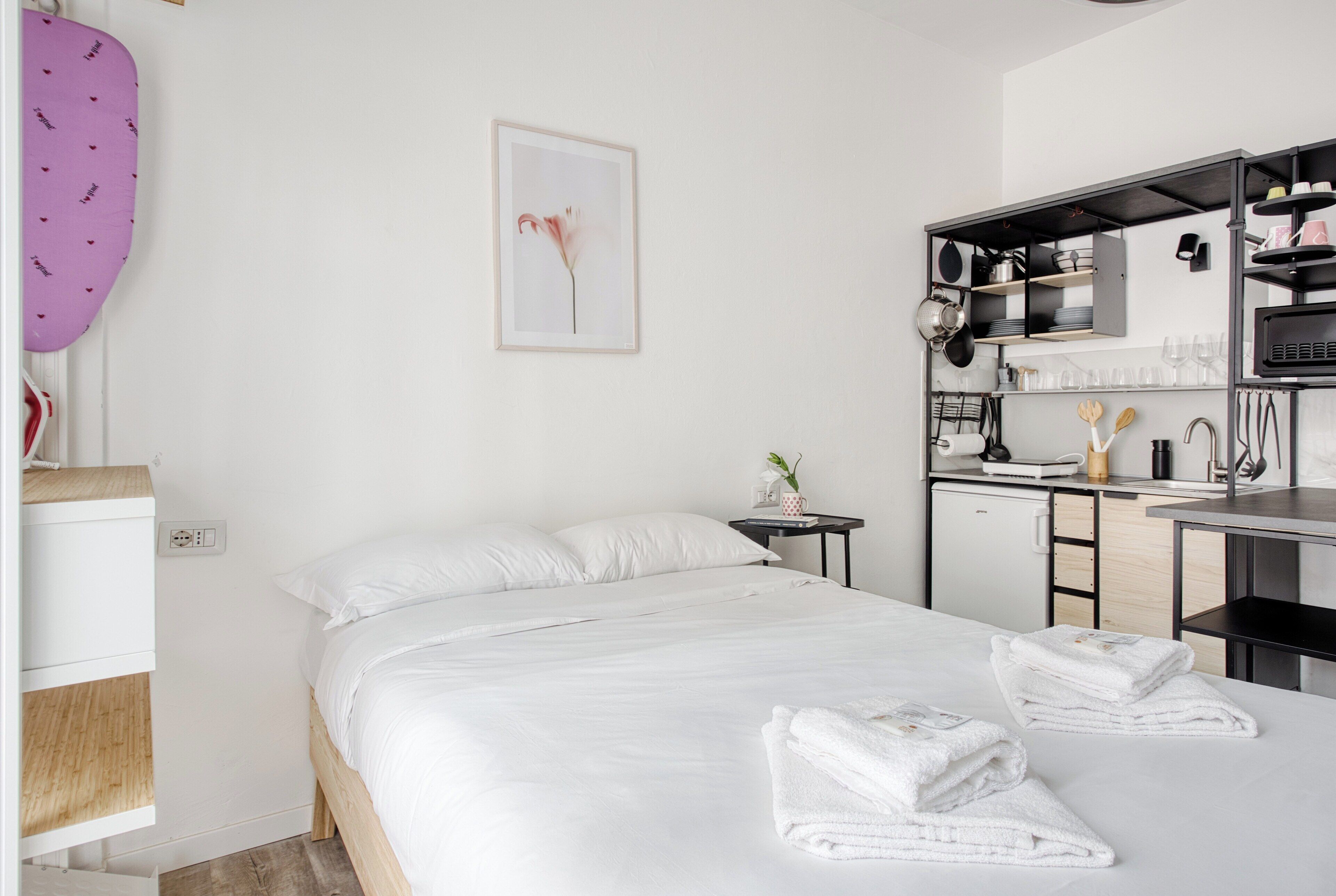 A modern hotel room with a neatly made bed, a small bedside table with a lamp, and a kitchenette area.