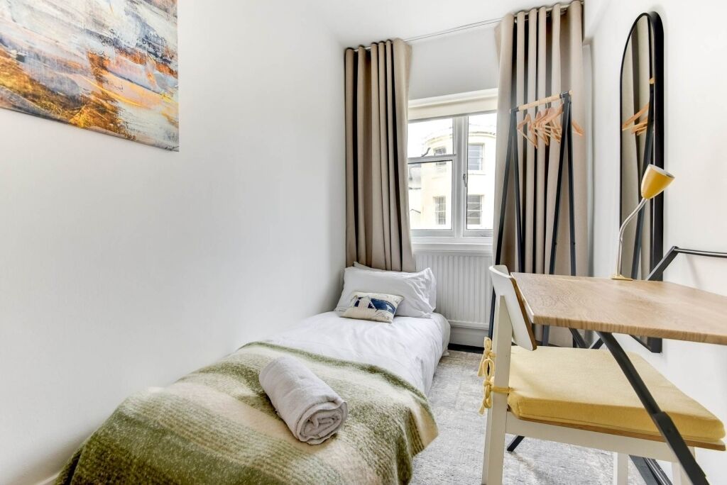 A small, neatly arranged bedroom with a bed, a chair, a table, and a window with curtains.