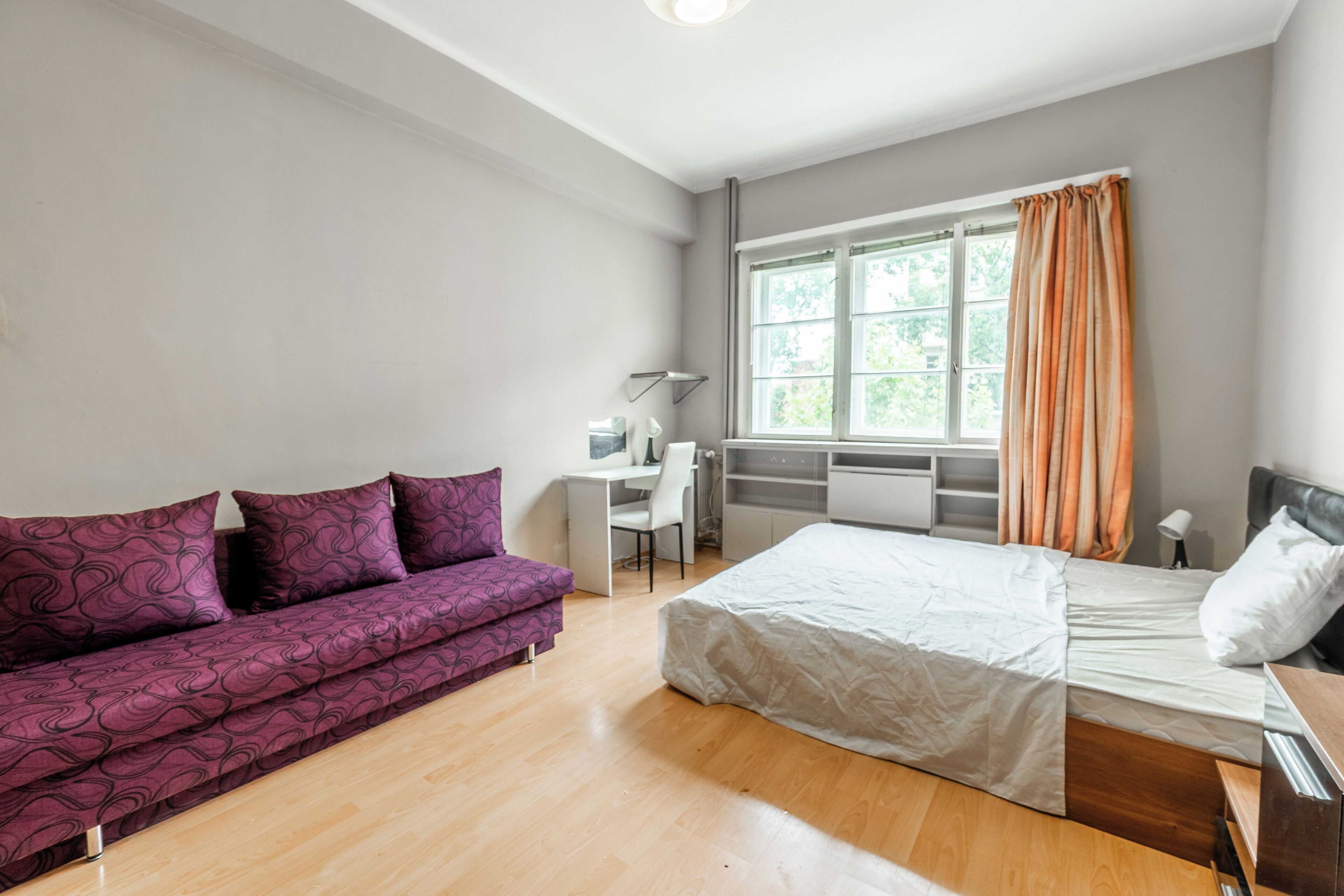 Un dormitorio moderno con cama, un sofá morado, un escritorio y una ventana con cortinas.