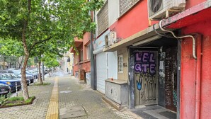 Una calle urbana estrecha con coches estacionados, una acera con pavimento táctil y un edificio con grafitis.