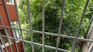 Un balcón con barandilla metálica, un edificio rojo y exuberantes árboles verdes al fondo.