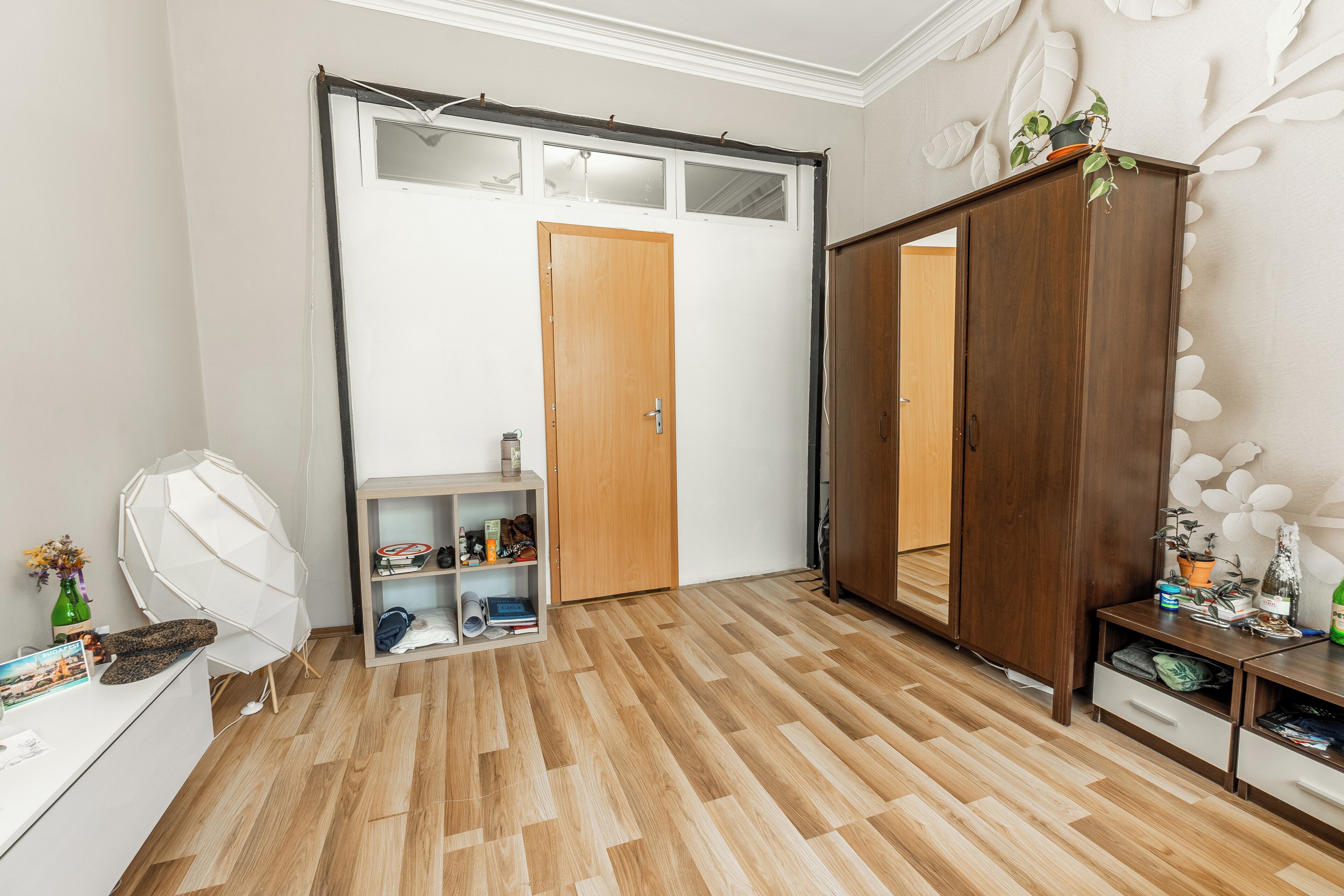 A room with a wooden floor, a wardrobe, a desk with a chair, and a shelf with various items.