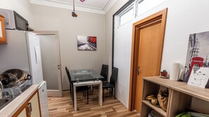 Una cocina compacta con refrigerador, microondas y una mesa de comedor con sillas. Hay una puerta que da a otra habitación y un estante con utensilios de cocina.