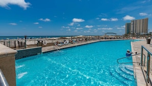A swimming pool with clear blue water, surrounded by lounge chairs and umbrellas, with a beach and ocean in the background. - Phoenix X 809 3 Bedroom Condo (Orange Beach)