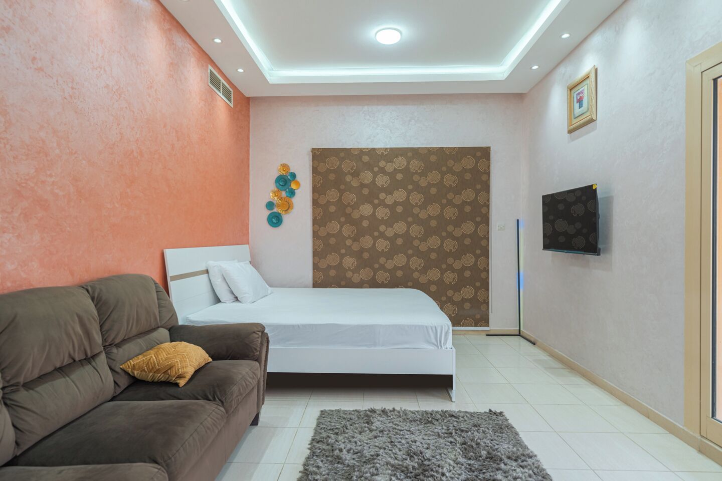 Une chambre à coucher comprenant un lit, un canapé, un tapis, un mur orné d’un panneau décoratif et un téléviseur fixé au mur.