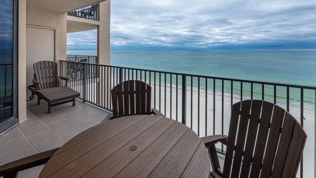 A balcony with a view of the ocean, featuring a wooden table and two Adirondack chairs.