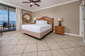 A hotel room with a large bed, a ceiling fan, a wooden nightstand with lamps, a round mirror, and a view of the ocean through a sliding glass door. - Phoenix X 303 3 Bedroom Condo (Orange Beach)