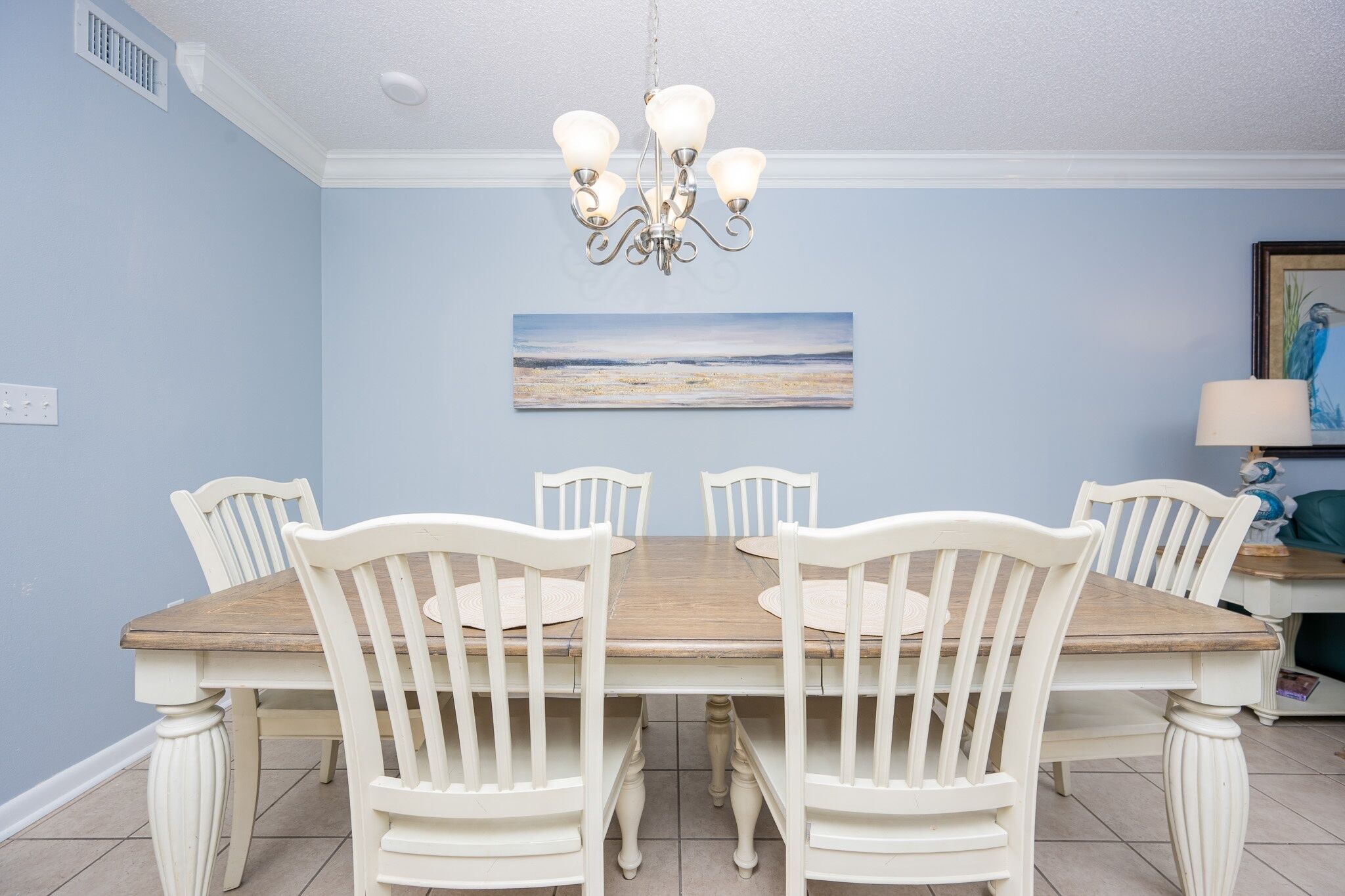 Un coin repas avec une table en bois et des chaises blanches, un lustre et un tableau représentant une plage accroché au mur.