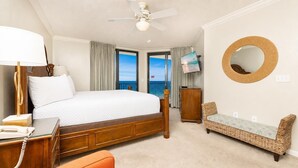 Une chambre d’hôtel avec un grand lit, un bureau avec un téléphone, une chaise, une télévision, un ventilateur de plafond et une vue sur l’océan.