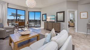 Een ruime woonkamer met een grote bank, een houten salontafel en een televisie. Er is een balkon met uitzicht op de oceaan.