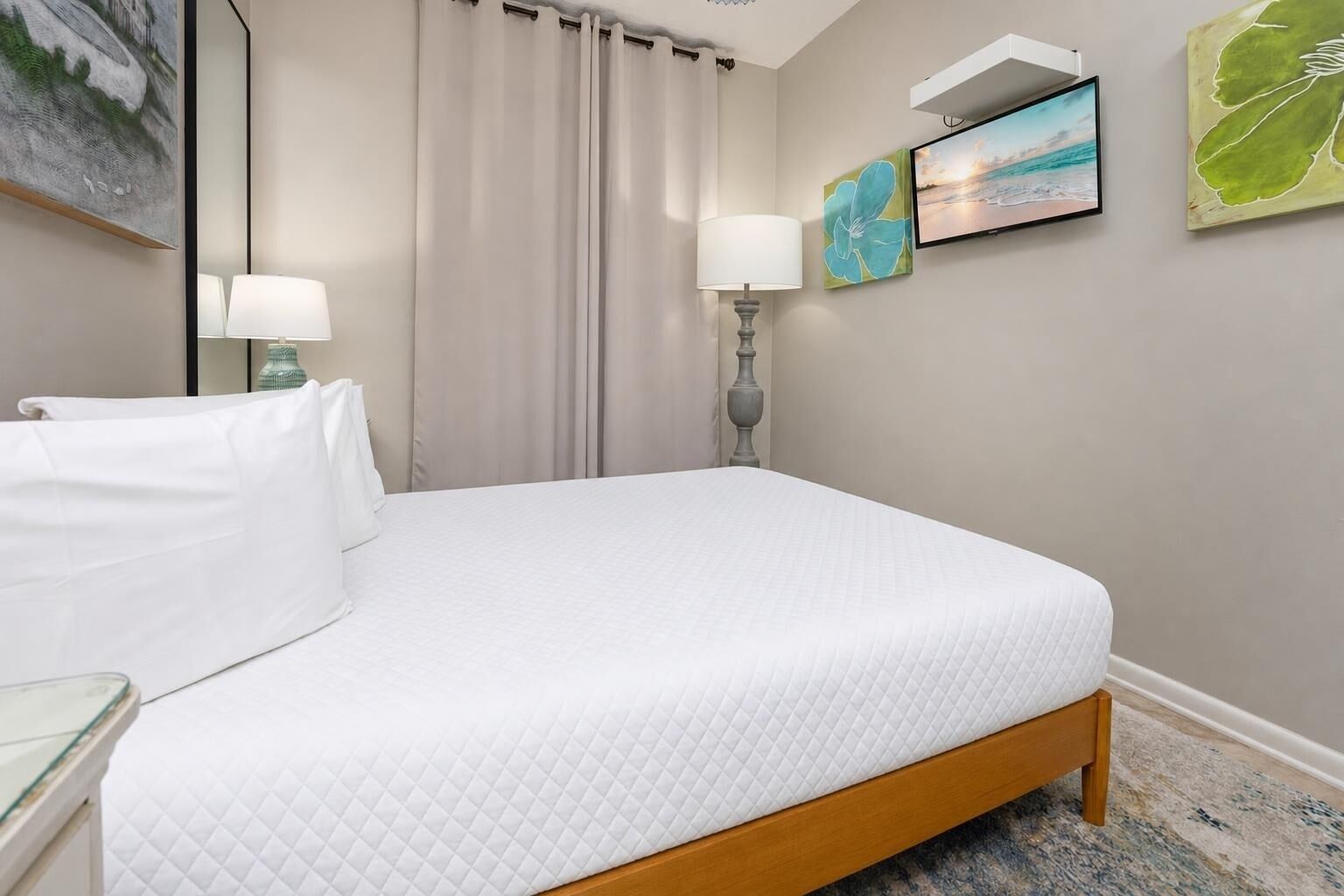 A neatly made bed with a quilted white cover, a bedside table with a lamp, and a wall with a painting and a framed picture.