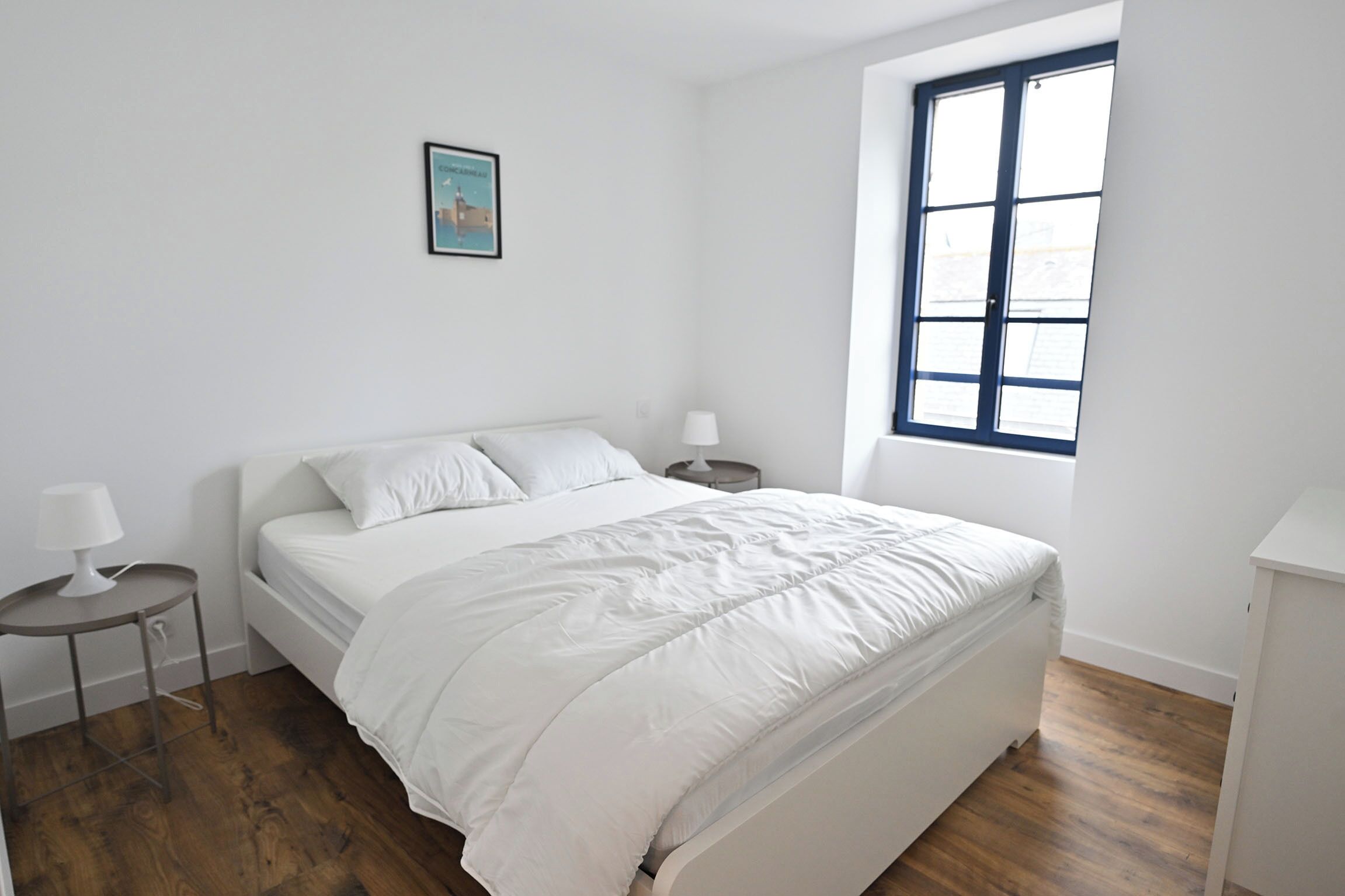 Une chambre à coucher avec un lit, une table de chevet, une lampe, une fenêtre et un tableau au mur.