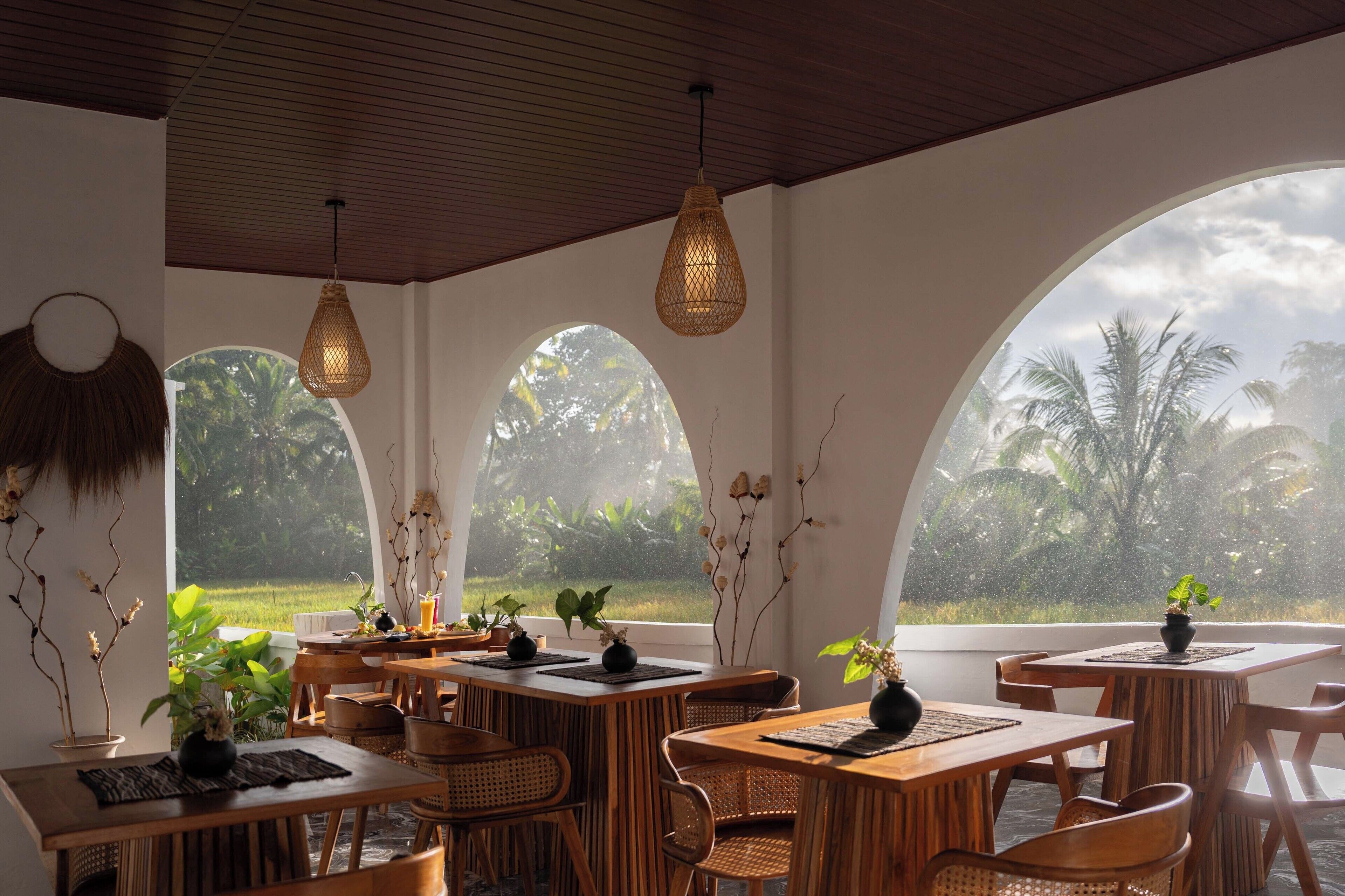 A restaurant with wooden tables and chairs, hanging lights, and large windows with views of greenery.