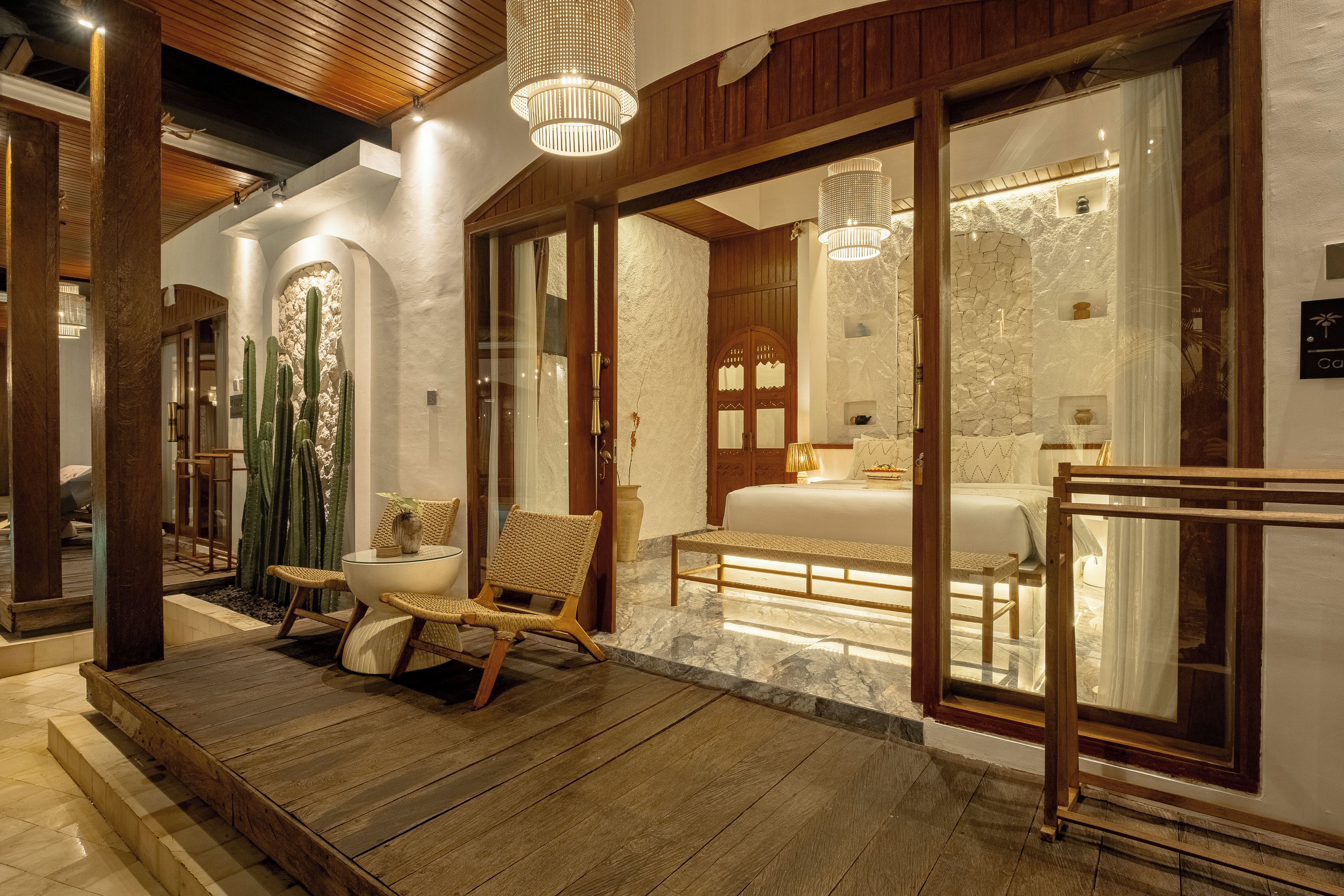 A hotel lobby with a wooden floor, a seating area with wicker chairs, a cactus display, and a large bed with a bathroom visible through a glass door.