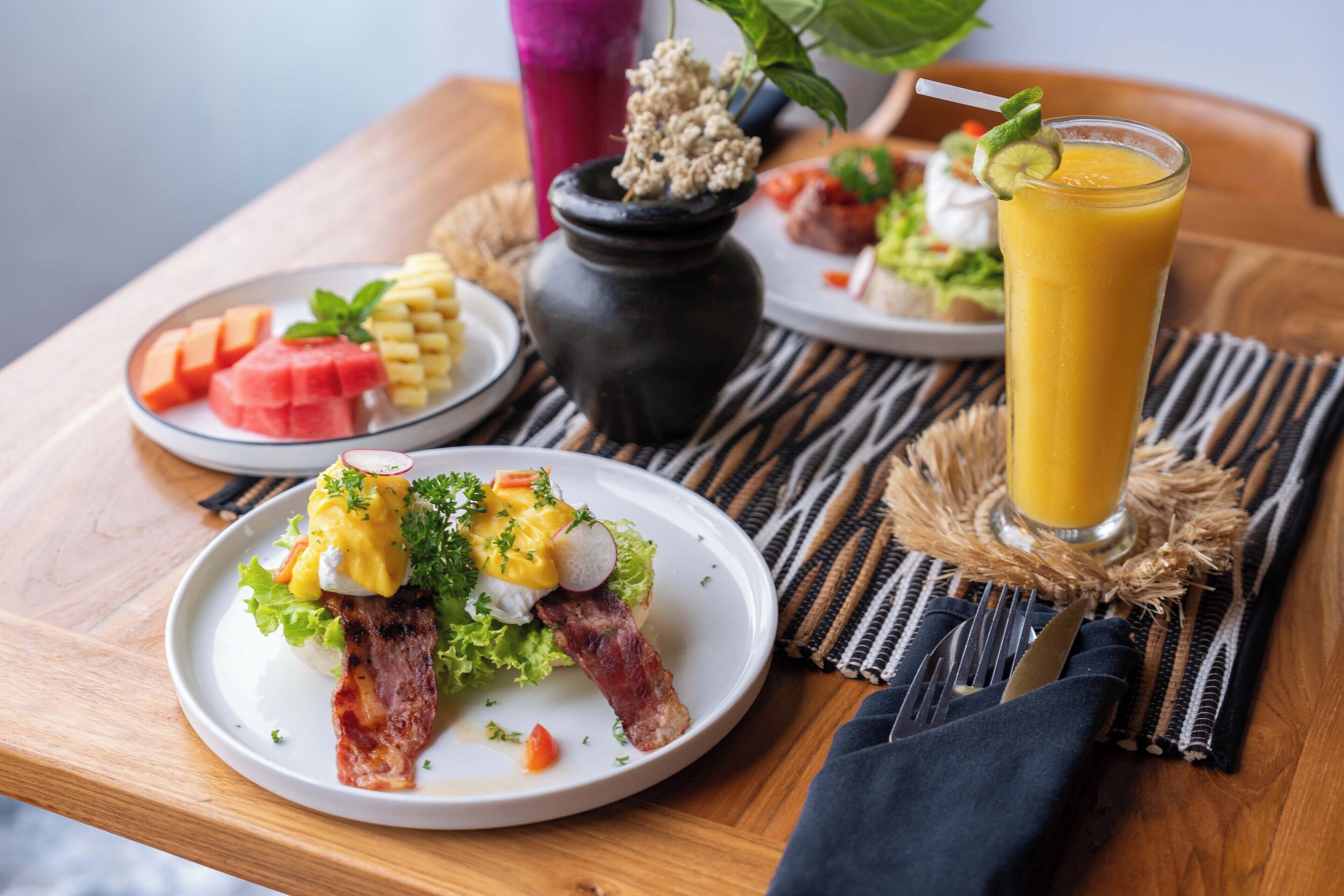 A breakfast setting with a plate of eggs, bacon, and vegetables, a glass of orange juice, and a small vase with dried flowers.