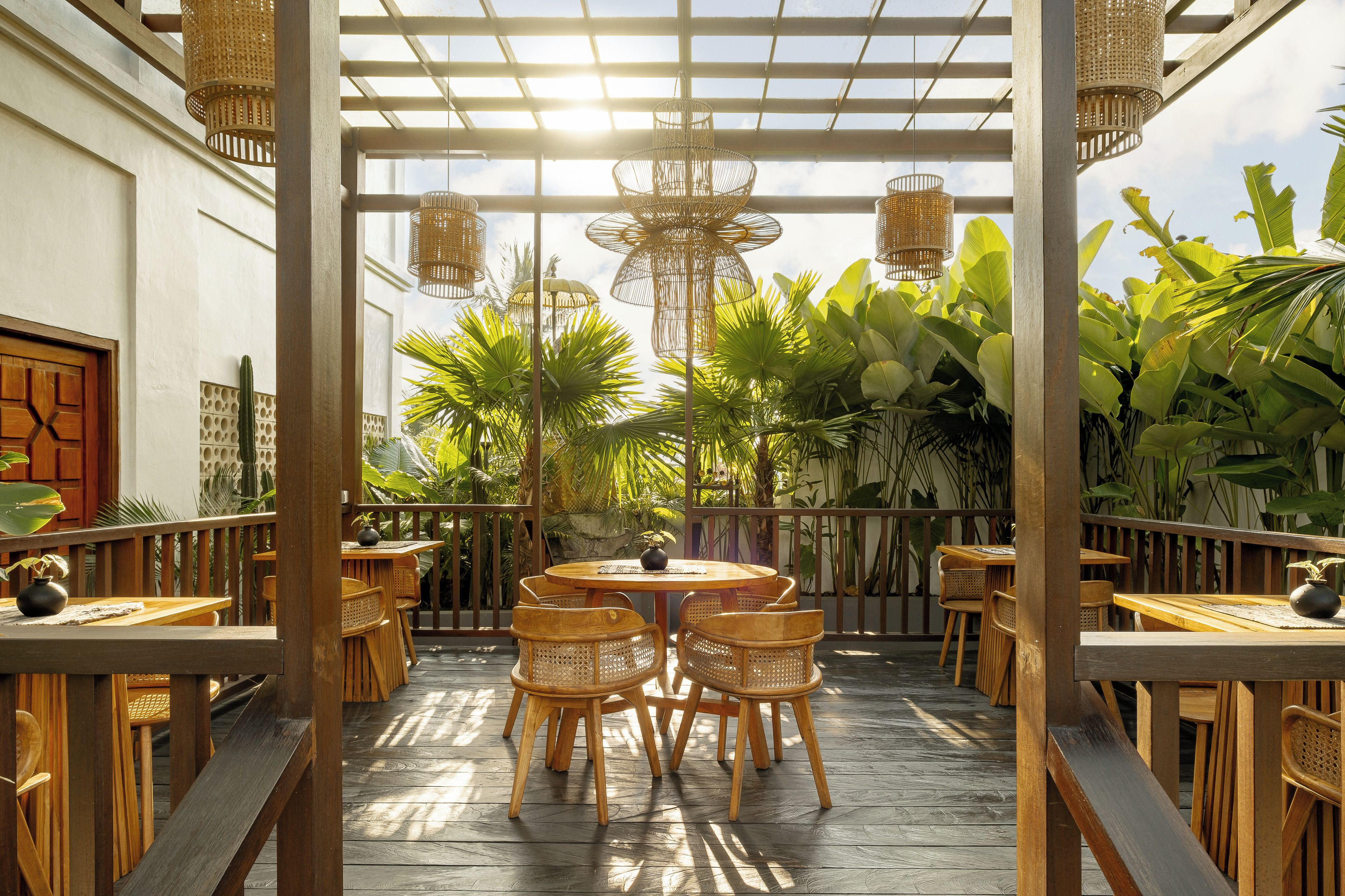 An outdoor patio with wooden tables and chairs, surrounded by lush tropical plants and a wooden pergola with hanging lights.
