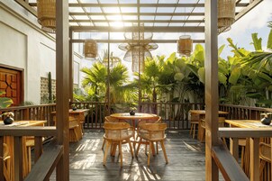 Un patio exterior con mesas y sillas de madera, rodeado de exuberante vegetación tropical y una pérgola de madera con luces colgantes.