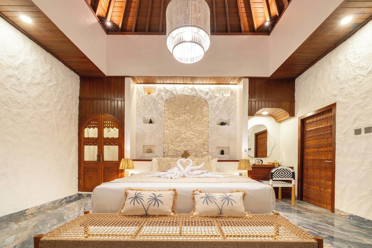 Um quarto espaçoso com uma cama grande, um redes de dormir e decoração rústica, uma parede de pedra, portas de madeira e um lustre.