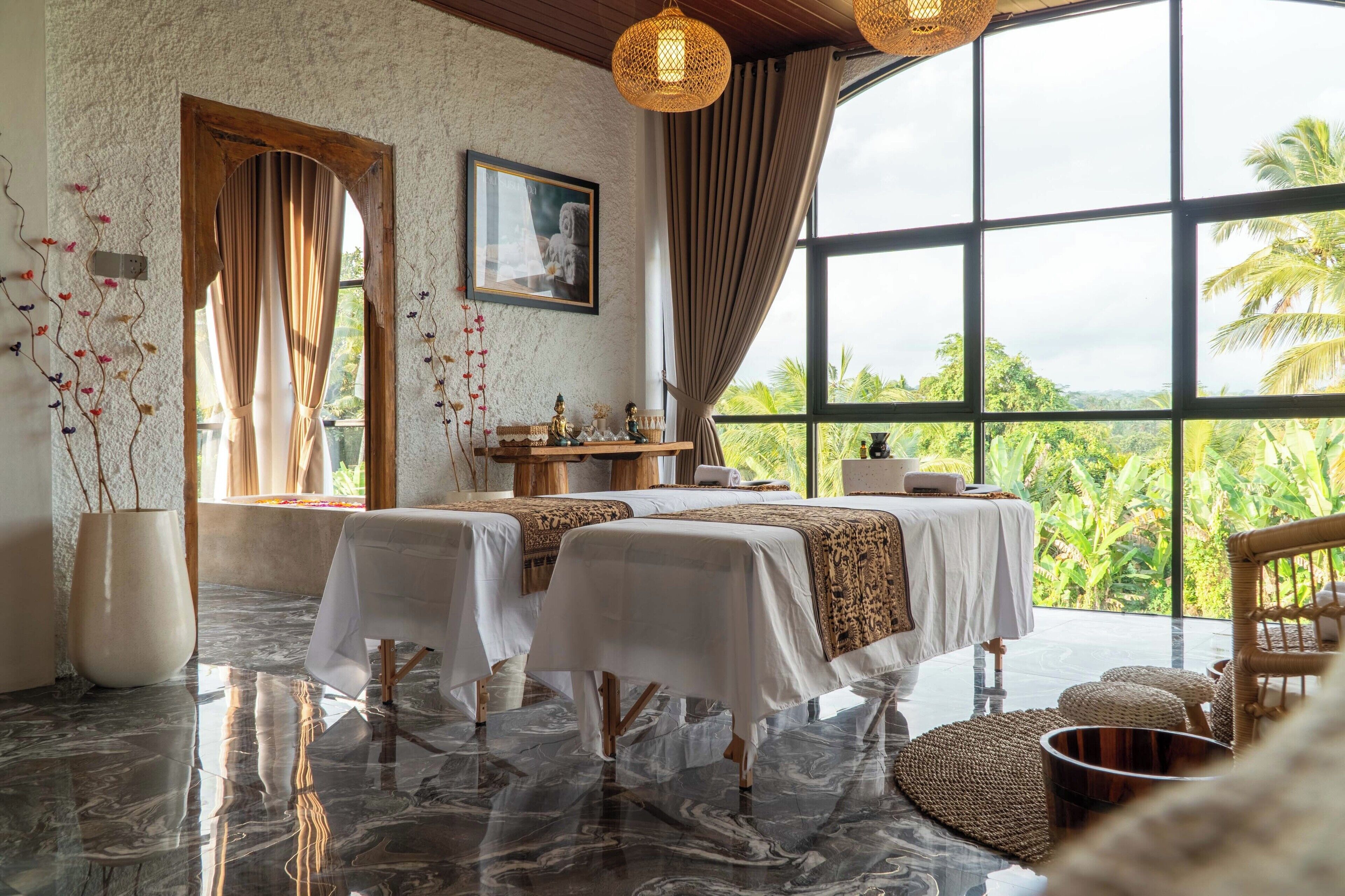 A room with two massage tables, a large window with curtains, a framed picture on the wall, and a view of greenery outside.