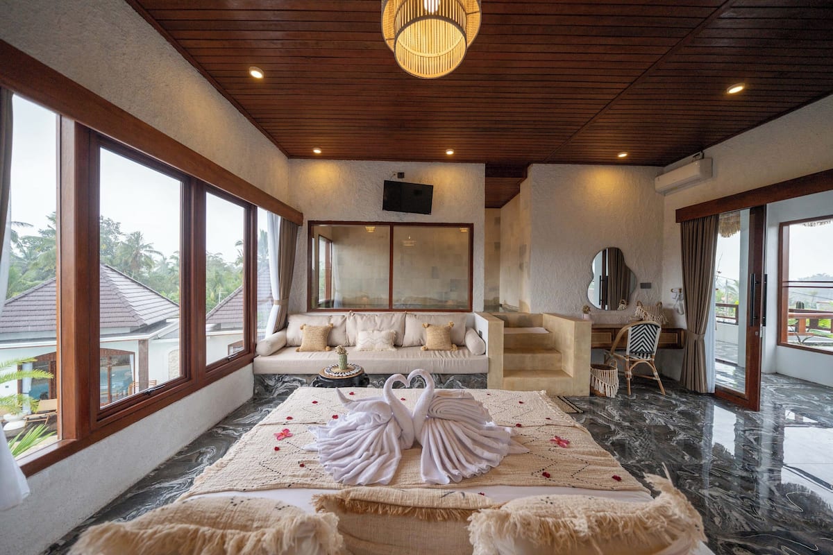 Quarto espaçoso com grandes janelas, teto de madeira, sofá, televisão e cama com toalhas dispostas em formato de cisne.