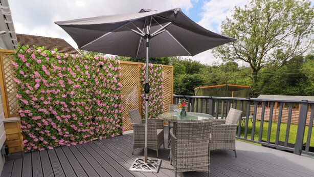 Una terraza con mobiliario de mimbre y una mesa de vidrio, rodeada por una valla de madera y plantas en flor.