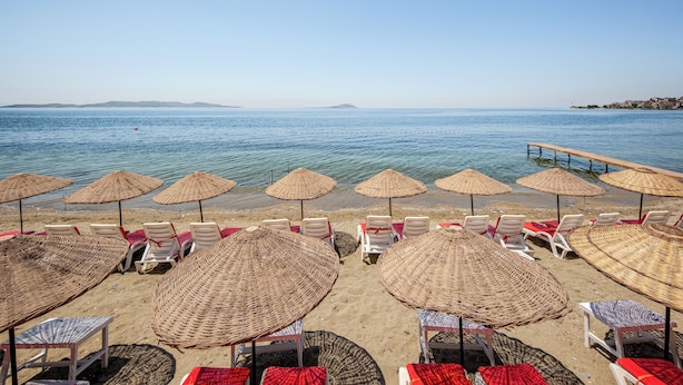 Ein Strand mit Reihen weißer Liegestühle und Strohdächern, ein Steg, der sich ins Wasser erstreckt, und freie Sicht auf das Meer und das entfernte Land.