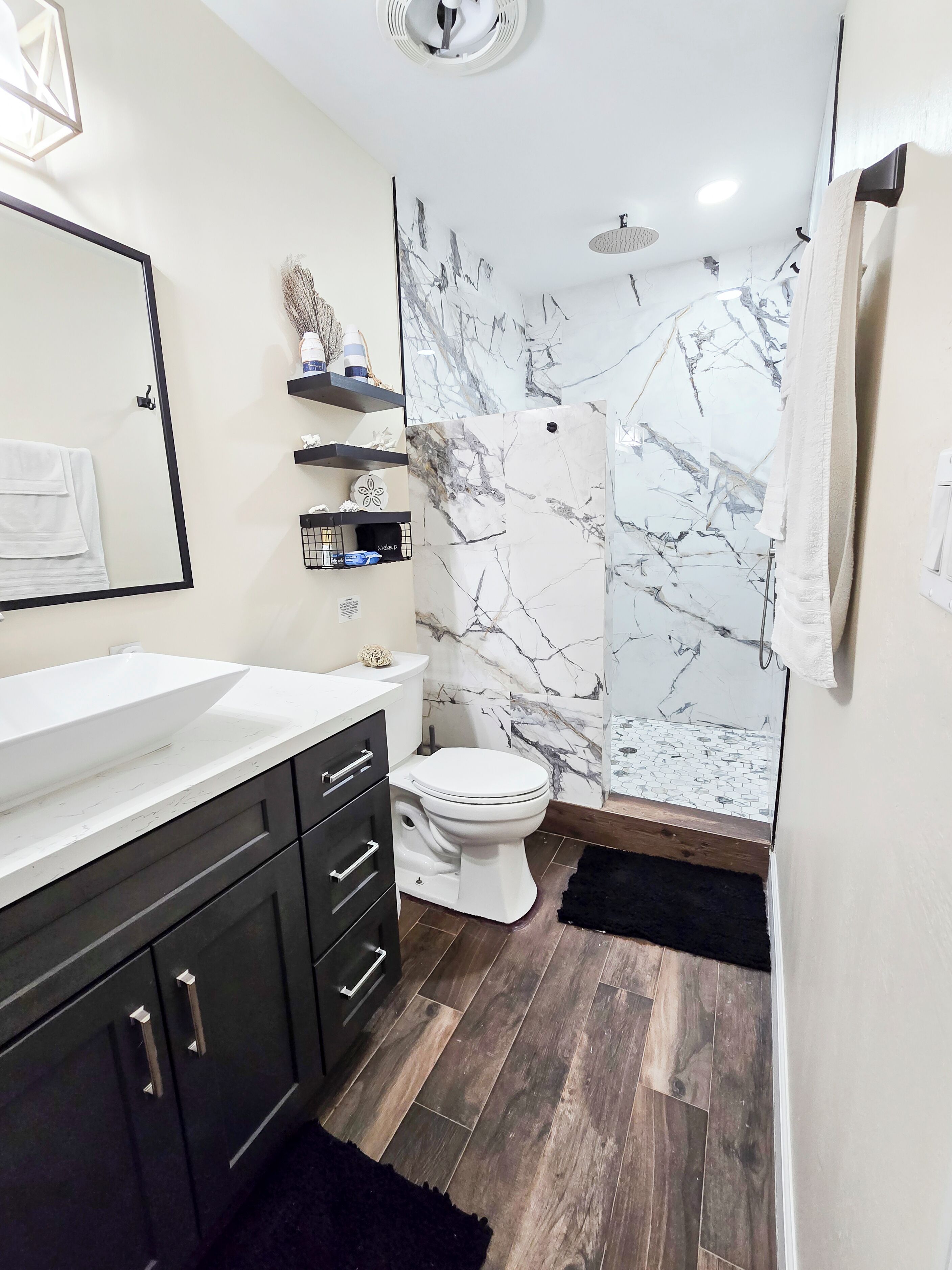 A modern bathroom with a white sink, dark cabinets, a toilet, and a shower with marble walls.