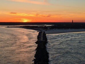 Eine Küstenlandschaft mit einem Steg, der ins Wasser ragt, einem Leuchtturm in der Ferne und einem Sonnenuntergang am Horizont.