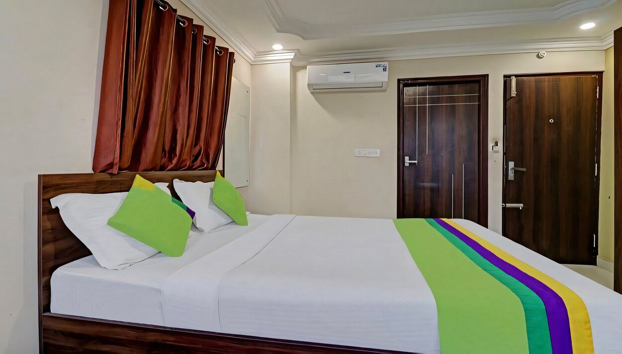 A hotel room with a bed, a wooden headboard, a white bedspread, green and white pillows, a striped blanket, a window with brown curtains, and a wall-mounted air conditioner.