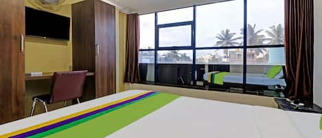 A hotel room with a large bed, a desk, a chair, a TV, and a window with a view of palm trees and buildings.