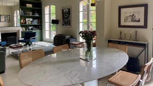A spacious living room with a marble dining table, comfortable seating, a large window, and a green bookshelf.