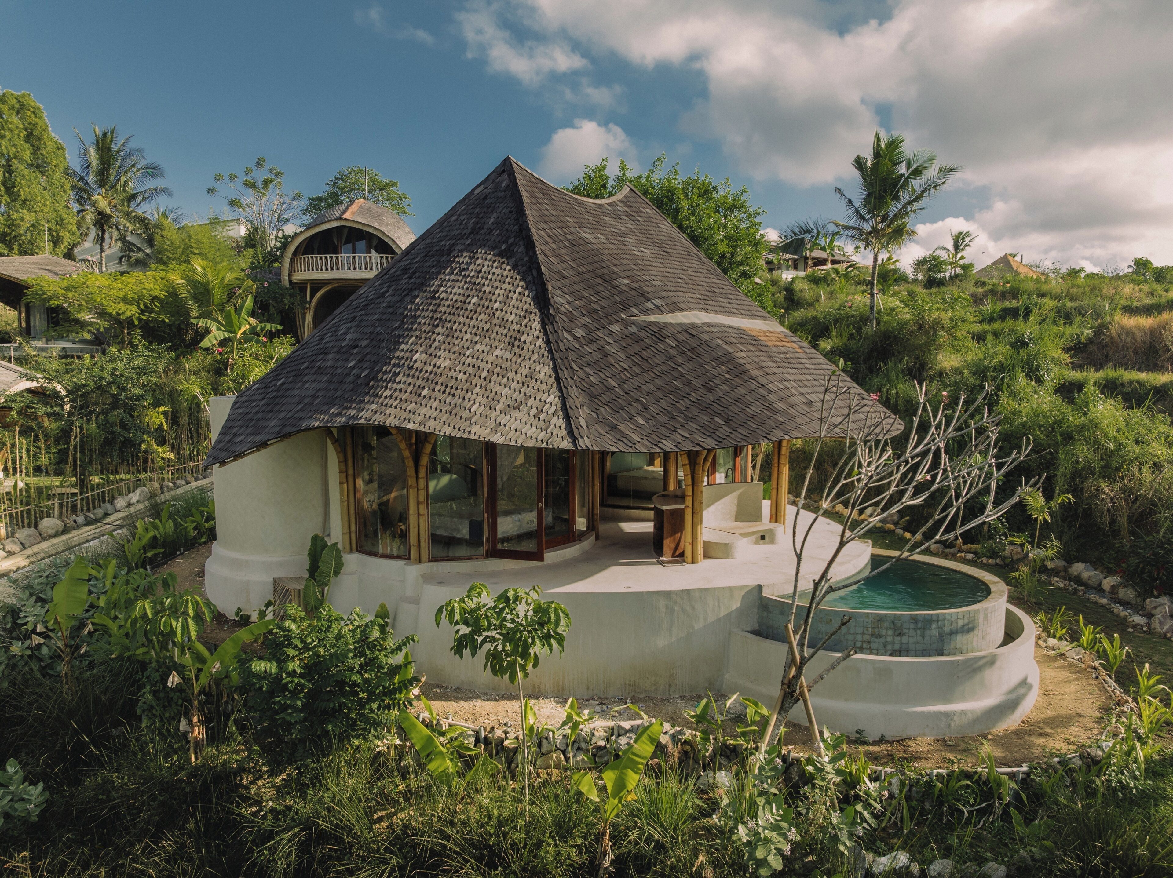 A bungalow with a thatched roof, surrounded by lush greenery and a swimming pool.