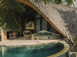 A pool area with a curved edge, a seating area with cushions, and a thatched roof structure. - MANDALA ECO RESORT (Selong Belanak)