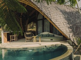 Un área de piscina con borde curvo, una zona de estar con cojines y una estructura de techo de paja.