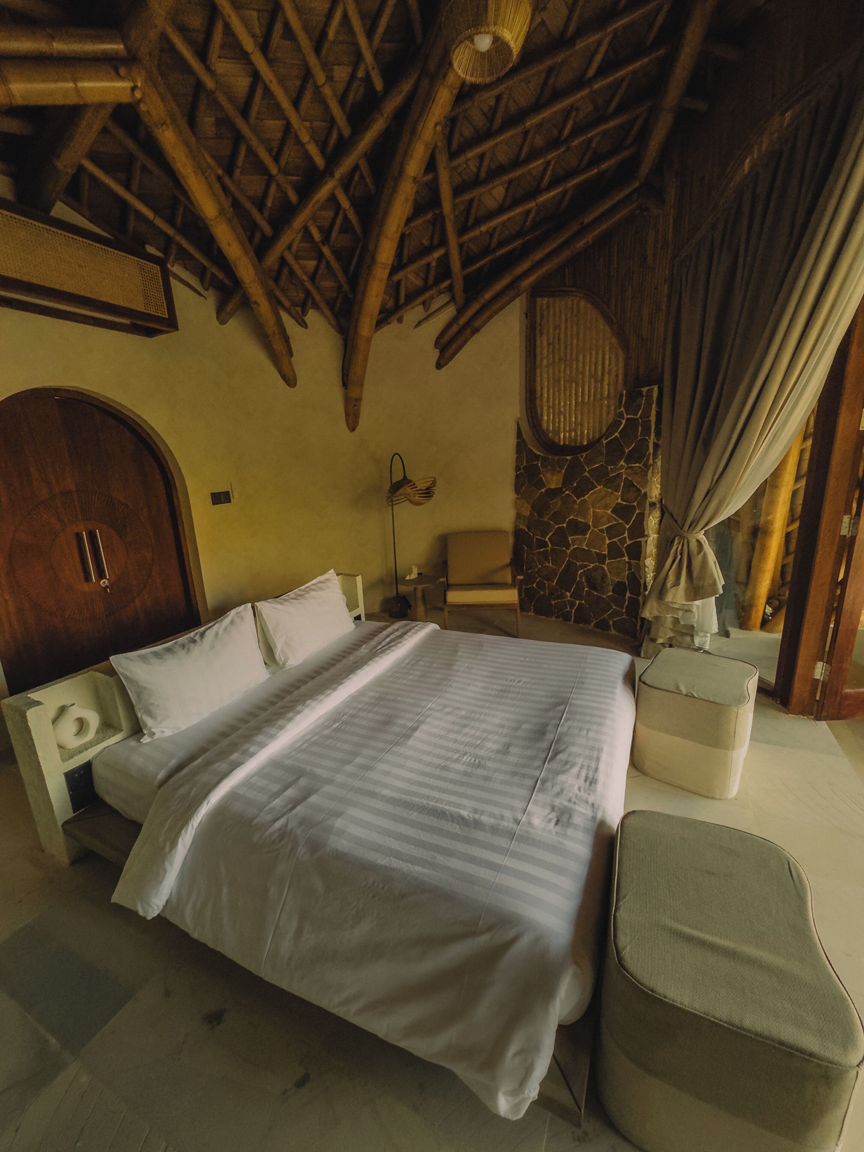 A room with a bed, a nightstand, a lamp, a stone wall, and a wooden ceiling.