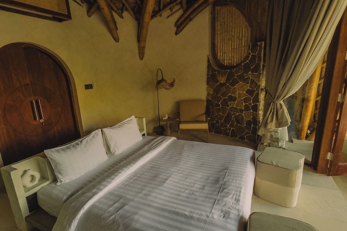 A room with a bed, a nightstand, a lamp, a stone wall, and a wooden ceiling.