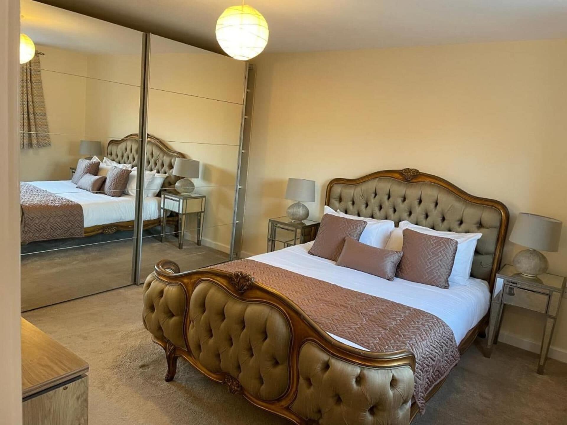 A bedroom with a large mirror, a tufted bed, bedside tables, and a lamp.
