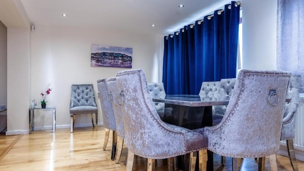 A dining area with a round table and four chairs, a blue curtain, and a painting on the wall. - 10 Guests - 5 Bedrooms - Free Parking - Lux House (Milton Keynes)