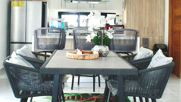A modern dining area with a rectangular table, six black wicker chairs, a chandelier, and a colorful rug.