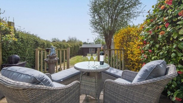 Un patio al aire libre con muebles de mimbre y una mesa dispuesta para dos personas, rodeado de exuberante follaje y vistas a una casa.