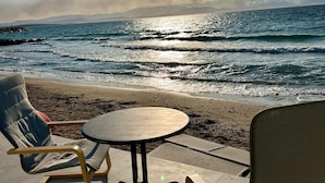 A beachside seating area with a table and chairs, overlooking the ocean with a clear view of the sun and distant mountains. - Aporte Hotel  (Çeşme)